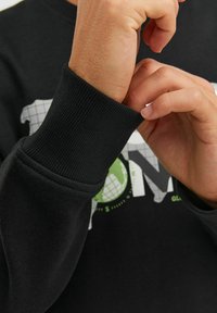 Sudadera negra con puños acanalados, que presenta un diseño gráfico impreso en blanco y verde en la parte frontal. Textura sutil evidente en la tela.