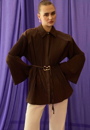 Femme aux cheveux plaqués en arrière portant une veste plissée marron foncé à manches larges, une ceinture, un pantalon blanc, et se tenant devant un rideau violet.