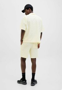 Camisa de manga corta amarilla-lima y pantalones cortos a juego con textura de patrón de olas. Una gorra negra y zapatos completan el atuendo informal.