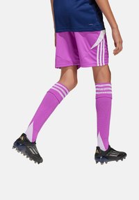Paarse voetbalkorte broek met witte accenten, gecombineerd met kniehoge paarse sokken met strepen en zwarte sportschoenen met zichtbare branding.