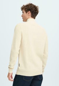 Crème gekleurde geribde trui met een hoge hals, lange mouwen en een getailleerde pasvorm, met een klein merkje aan de zijkant.
