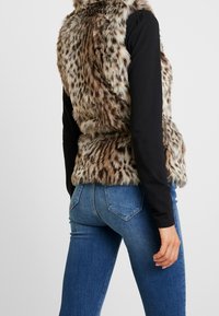 Faux-Fur-Weste mit einem gepunkteten Muster in Braun und Creme, die eine weiche Textur aufweist. Getragen über einem schwarzen Langarmhemd und blauen Jeans.