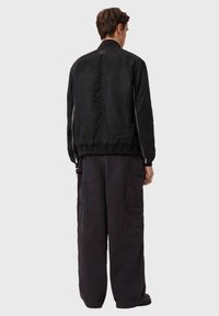 Μαύρο μπουφάν bomber με λεία υφή, ελαστική τρέσσα στο κάτω μέρος και τσέπες με φερμουάρ, συνδυασμένο με χαλαρά σκούρα cargo παντελόνια.