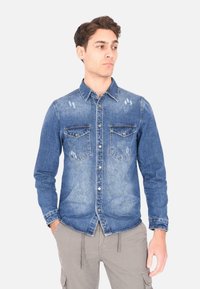 Camicia in denim blu con colletto classico, due tasche sul petto e dettagli strappati. Maniche lunghe con polsini abbottonati e orlo diritto.