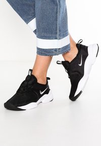 Svart atletiska skor i mesh-material med en vit sula. Har en silverfärgad logga och svarta snörningar. Bärs med uppvikt denimjeans.