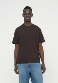 Καφέ βαμβακερό T-shirt με κοντά μανίκια και στρογγυλή λαιμόκοψη, με μικρό, σκούρο λογότυπο στο στήθος. Συνδυασμένο με ανοιχτό μπλε τζιν παντελόνι.