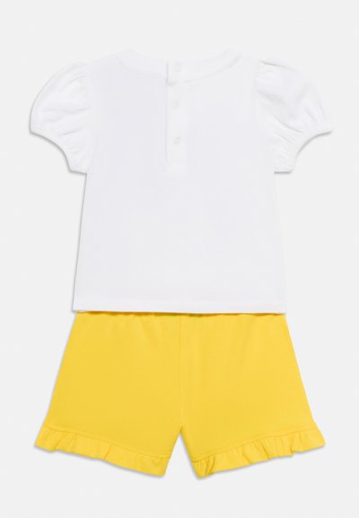 Λευκό τοπ με κοντά μανίκια και κούμπωμα με κουμπιά στο πίσω μέρος, συνδυασμένο με κίτρινα σορτς με φαρδιά τελειώματα και μαλακή υφή.