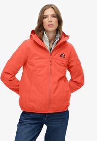 Kvinde iført en rød hættetrøje med lynlås over en stribet skjorte og blå jeans, stående med hænderne bag ryggen på en hvid baggrund.