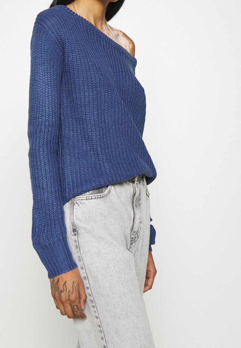Pull bleu tricoté avec un col asymétrique, une texture côtelée et des manches longues, associé à un jean taille haute gris clair.