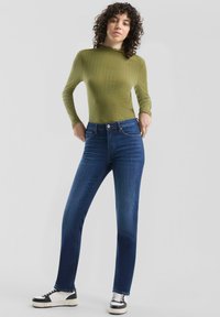 Długi top w kolorze oliwkowym z prążkowanego materiału, zestawiony z ciemnoniebieskimi jeansami o prostych nogawkach oraz czarno-białymi trampkami, z dopasowanym krojem.