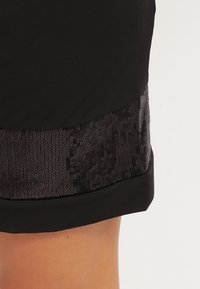 Svarta shorts med en slät överdel av tyg och en nedre band i svarta paljetter. Texturen av paljetterna tillför ett glansigt inslag till designen.