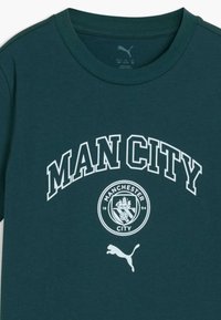 T-shirt vert foncé à col rond avec texte blanc "MAN CITY", logo du club de football de Manchester City et logo blanc Puma en dessous du logo.