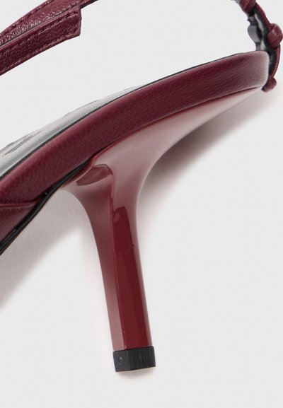 Izbliza prikaz tamnocrvene štikle s fokusom na peta i kaiš oko gležnja na bijeloj pozadini.