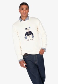 Jersey crema con gráfico de un perro azul y el texto "Hugo & Nora", combinado con jeans de mezclilla azul oscuro y un cuello de camisa a cuadros.