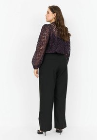 Lace bluse i mørk lilla med et blomstermønster, lange blazede ærmer og en tætsiddende ryg, parret med højtaljede sorte bukser.