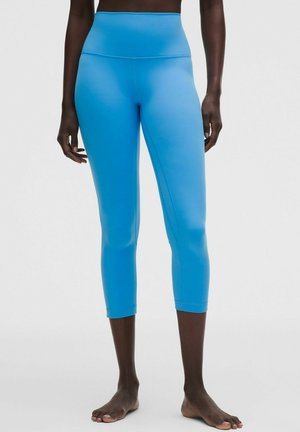 Donna con leggings corti vita alta di colore blu acceso, in piedi a piedi nudi su uno sfondo chiaro uniforme.
