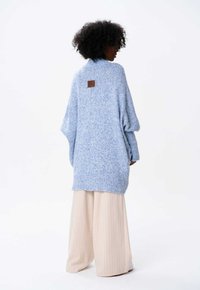  Lichtblauwe gebreide oversized trui met een hoge kraag en ribbelrand. Bevat een bruine leren patch op de achterkant. Gecombineerd met beige gestreepte broek.