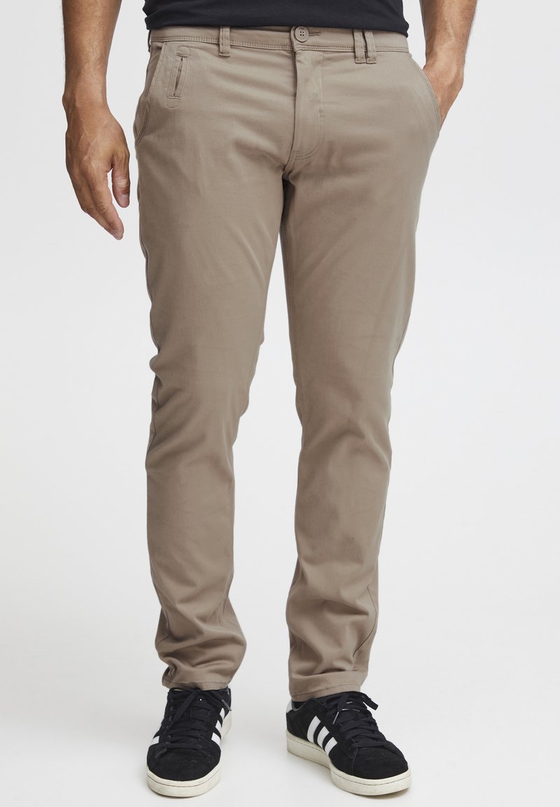 Pantalons en coton beige avec une coupe slim, dotés de poches latérales et d'une fermeture à boutons, associés à des baskets noires avec des accents blancs.