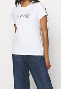 Mujer vistiendo una camiseta blanca de Levi's con letras florales y vaqueros azules, de pie frente a un fondo blanco liso.