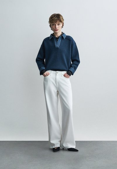 Ένα άτομο στέκεται με τα χέρια στις τσέπες, φοράει σκούρο μπλε πουλόβερ με γιακά και μακριά μανίκια, λευκό παντελόνι φαρδύ και μαύρα παπούτσια με μυτερή μύτη, σε γκρι πάτωμα.