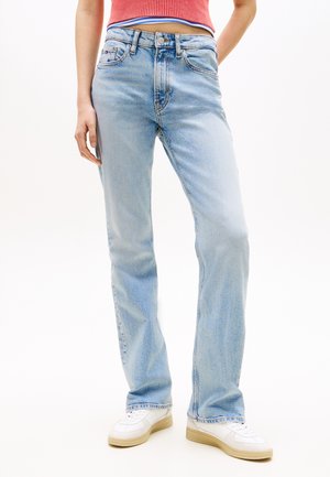 Person iført lyseblå, lige ben jeans, hvide sneakers og en cropped rød ærmeløs top mod en hvid baggrund.