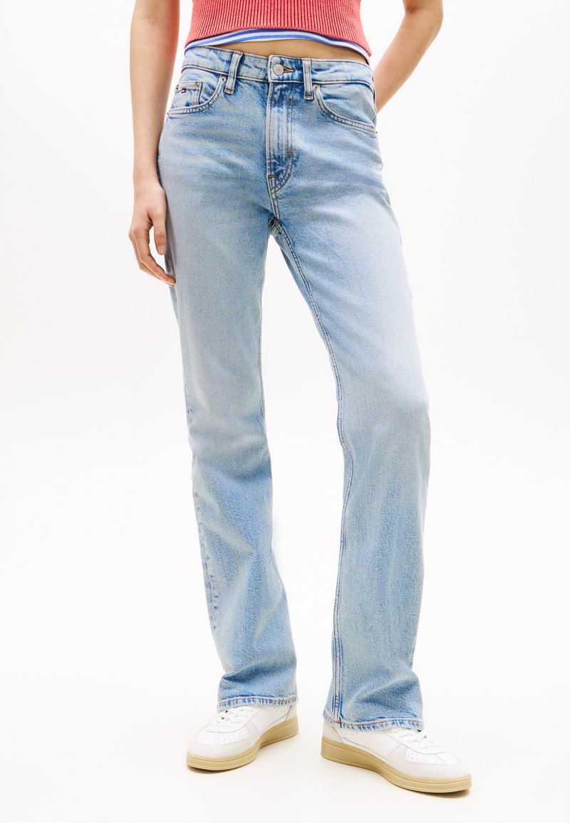 Άτομο που φοράει ανοιχτό μπλε τζιν παντελόνι με ίσια γραμμή, λευκά αθλητικά παπούτσια και κόκκινο κόψιμο αμάνικο τοπ, μπροστά σε λευκό φόντο.