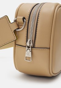 Borsa in pelle beige con texture liscia, dotata di una cerniera metallica, di un tirazip con logo e di un'etichetta gialla a contrasto.