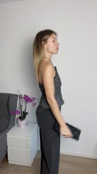 Schwarzes Peplum-Top mit dünnen Trägern, kombiniert mit einer schwarzen Hose. Hält eine strukturierte schwarze Clutch. Der Hintergrund zeigt ein graues Sofa und topfgepflanzte Orchideen.