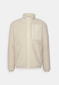 Beige fleecejacka med hög krage, dragkedja framtill och en ficka med lappar på bröstet. Har sidofickor med dragkedja och ribbade ärmslut.