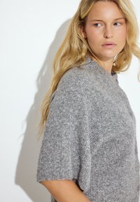 Pilka trumpa rankovėmis megztinis, pagamintas iš tekstūruoto mezgimo audinio su aukštu kaklu. Turi laisvą siluetą ir subtilius raukšlėjimus, suteikiančius papildomo žavesio.