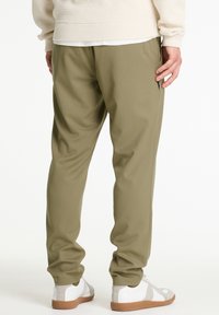 Olívazöld elasztikus nadrág sima textúrával, keskenyedő fazonban, oldalzsebekkel, mögülnézetből látható fehér cipőkkel, szürke részletekkel.