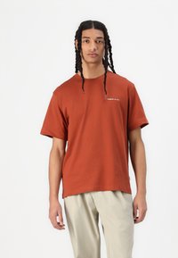 Tricou din bumbac de culoare ruginie, cu mâneci scurte, având un mic logo alb pe piept. Modelul poartă pantaloni ușori de culoare bej.