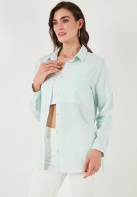 Light mint green button-up shirt with a pocket, made of textured fabric; features a collar and full-length sleeves, slightly frayed at the hem.