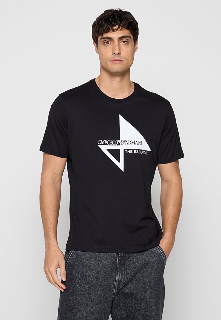 Hombre que lleva una camiseta negra de manga corta con el logo de Emporio Armani y el texto "The Essence", combinada con vaqueros gris oscuro, de pie con una mano en el bolsillo.
