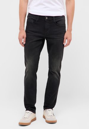 Mann trägt ausgewaschene schwarze Jeans und weiße Sneakers, steht vor einem schlichten hellen Hintergrund.