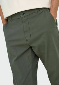 Hombre con pantalones verde oliva y manos en los bolsillos laterales, vistiendo una camisa blanca metida por dentro.