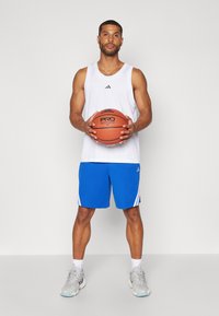 Fehér ujjatlan póló fekete logóval, kék rövidnadrág fehér csíkokkal, narancssárga kosárlabdát tart, szürke tornacipőt visel kék díszítéssel.