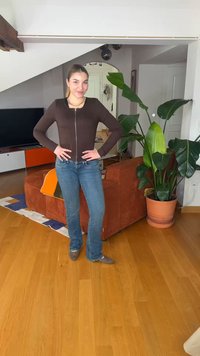 Γυναίκα που χαμογελά με τα χέρια στη μέση, φορώντας καφέ ζακέτα και μπλε τζιν, στέκεται σε σαλόνι με πορτοκαλί καναπέ και μεγάλο γλαστρόφυτο.