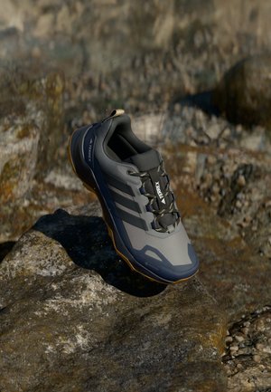 Grå og sort Adidas Terrex vandresko på klippeoverflade, der viser side og top med snørebånd og rillede såler.