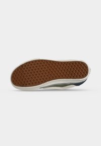 Semelle de chaussure avec une bande de roulement en caoutchouc brun à motif gaufré, une semelle intermédiaire blanche et des parties visibles bleues et vertes de la tige.