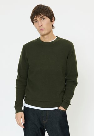 Mand iført mørkegrøn strikket trøje og mørke jeans, stående med den ene hånd i lommen mod en ensfarvet hvid baggrund.