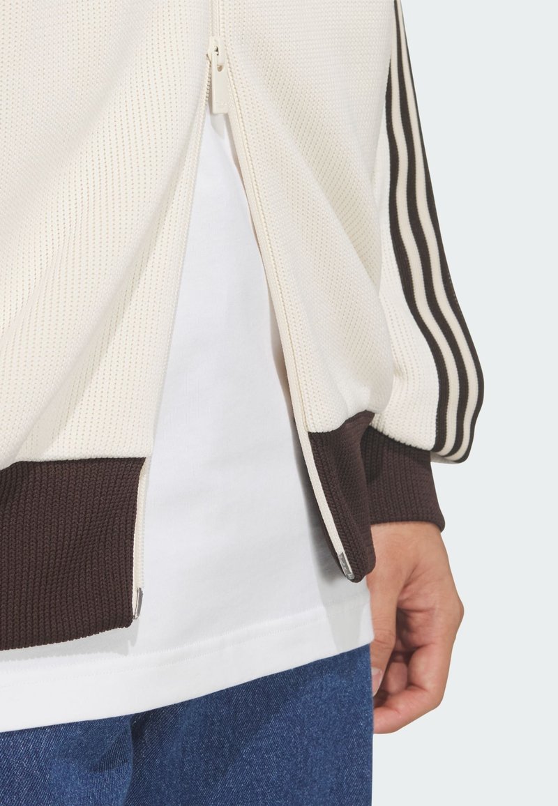 Gedeeltelijk zicht op een persoon die een roomkleurige trainingsjas met halve ritssluiting, bruine boorden en strepen draagt, een wit shirt en blauwe spijkerbroek, hand ontspannen langs de zijkant.