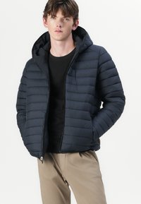 Marineblau wattierte Jacke mit Kapuze, ausgestattet mit einer gesteppten Textur und einem Frontreißverschluss. Kombiniert mit einem schwarzen Oberteil und hellfarbenen Hosen.
