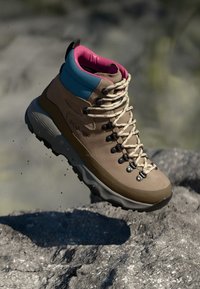Brown hiking boot with a textured surface, synthetic upper, pink and blue accents, and beige laces. Rugged sole designed for traction.
