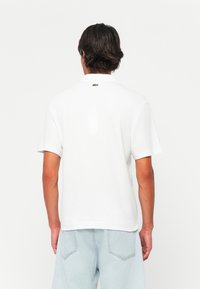 Polo shirt blanc à manches courtes, fait en coton. Présente un logo vert dans le dos. Conception simple, coupe standard et texture lisse.