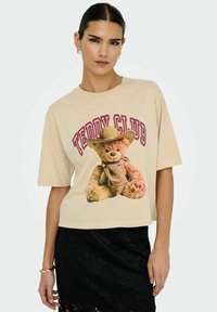 Beige cropped t-shirt van katoen, met een teddybeerafbeelding in een cowboyhoed en de tekst "Teddy Club" in bordeauxrood. Korte mouwen.