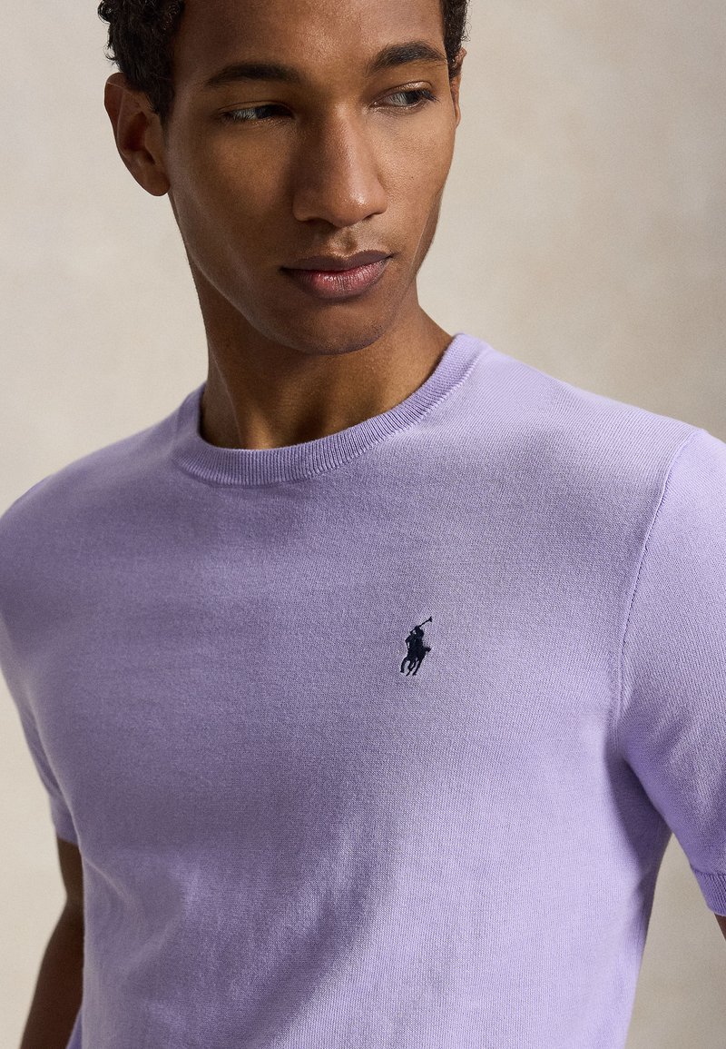 Jeune homme portant un t-shirt violet clair à col rond avec un petit logo brodé sombre d’un joueur de polo sur la poitrine.