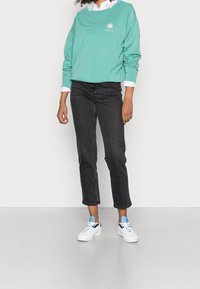 Felpa verde menta con logo del sole, camicia bianca con colletto sotto, jeans neri a vita alta e sneakers bianche con accenti blu.