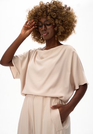 Czarnoskóra kobieta o kręconych włosach, nosząca duże okulary oraz luźną, beżową bluzkę i spodnie, poprawiająca okulary jedną ręką, druga ręka w kieszeni.