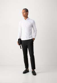Camisa branca de mangas compridas com botões, calças pretas ajustadas e mocassins pretos. A pessoa segura uma pequena bolsa preta. Fundo simples.